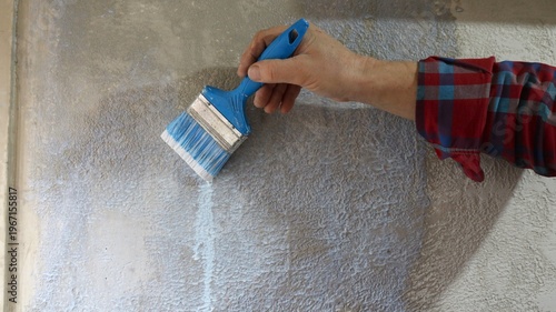 Close up of man in plaid shirt holding blue paintbrush applying liquid primer to textured grey plaster wall and priming wall of house with uneven textured plaster using paintbrush.