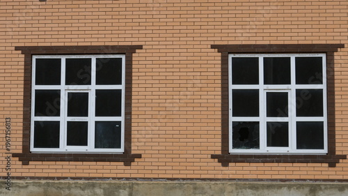 Two large windows with plastic frames and crossbars and broken glass from a thrown stone in the structure of a light brick house, the concept of danger and crime in residential areas