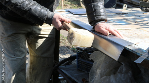 A craftsman uses a mallet to strike a bent sheet of iron while holding the material to a workbench, using a special hammer to strike a flexible piece of metal to secure the bend.