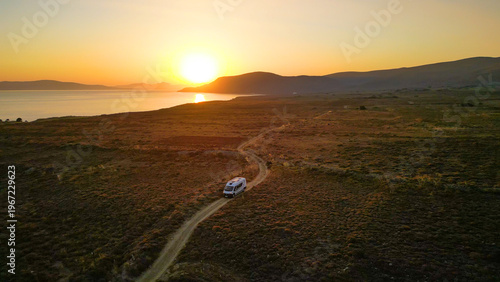 A camper van driving along a remote coastal road at sunset on Limnos Island, Greece. Aerial perspective shows golden Mediterranean light, rugged landscape and freedom of van life travel.