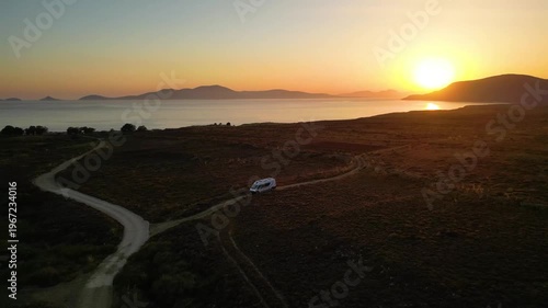 A camper van driving along a remote coastal road at sunset on Limnos Island, Greece. Aerial perspective shows golden Mediterranean light, rugged landscape and freedom of van life travel.
