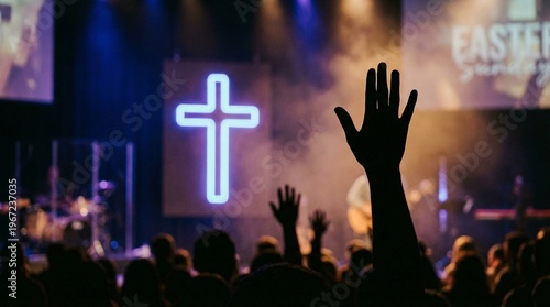 Silhouetted hand raised in worship before a glowing neon cross. Christian concert prayer service at church. Spiritual praise and adoration during Easter Sunday worship celebration. 