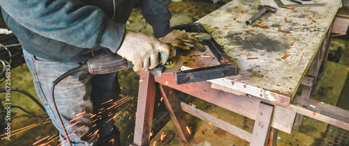 Male worker wearing protective gloves using an angle grinder to cut and shape a metal object on a workbench, generating a shower of bright orange sparks.