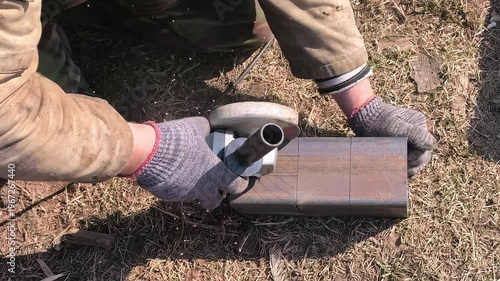 A person is focused on cutting a metal piece with a tool. The person wears gloves and is positioned on the ground. There is grass and dirt around the area.