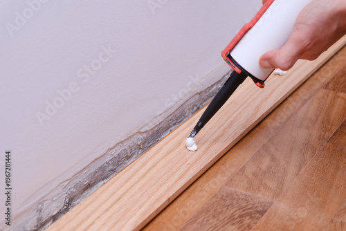 Hand applying white adhesive along a baseboard with a caulking gun