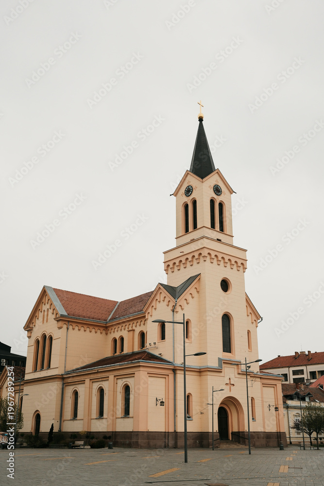Fototapeta premium Church building with tower located in Zrenjanin, Serbia under cloudy sky. Religious architecture and European city landmark.