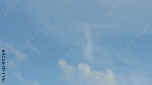 A rocket climbs high into a blue sky surrounded by soft clouds with a bright flame trail visible below. The spacecraft moves upward through the atmosphere leaving a glowing streak against the open sky