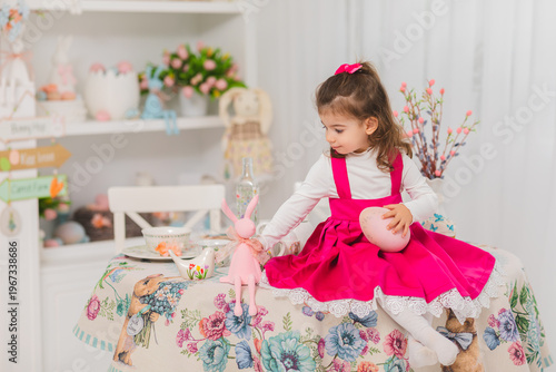 little girl in pink dress sitting on festive table holding easter egg surrounded by spring decor, toys and flowers in bright cozy home interior