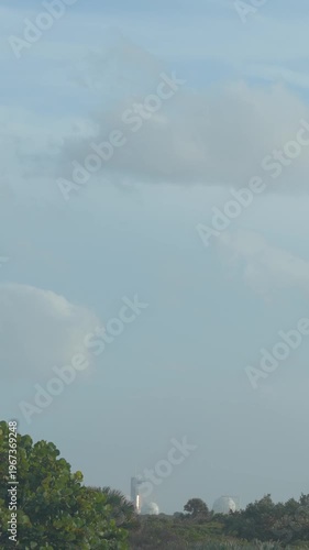 A rocket launches vertically from a coastal space center with trees and infrastructure visible below. Bright engine flames rise into a cloudy blue sky as the spacecraft lifts off from the launchpad.