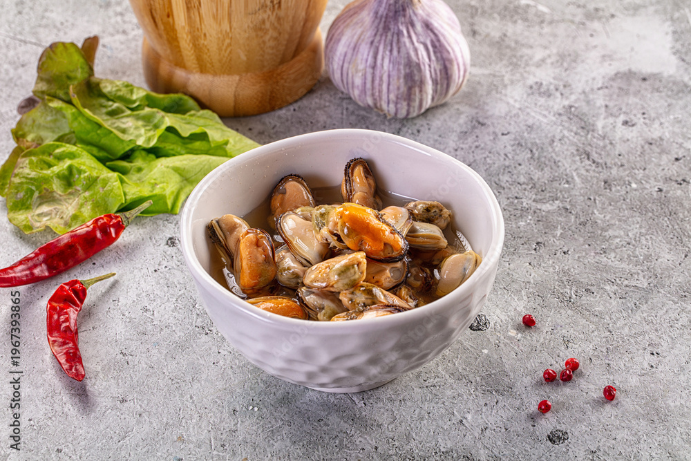Fototapeta premium Marinated mussels in a white bowl with chili peppers, garlic, and fresh lettuce on a concrete background.