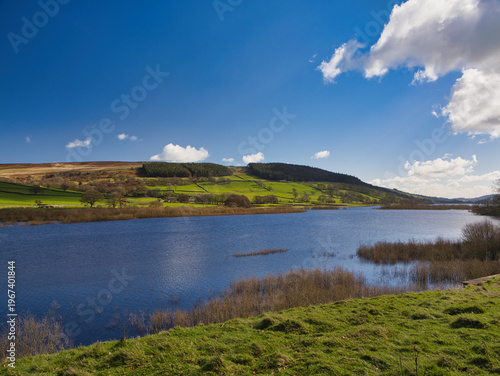 Bright view of Gouthwaite Reservoir in Yorkshire UK with blue water rolling hills and scattered clouds in crisp spring light