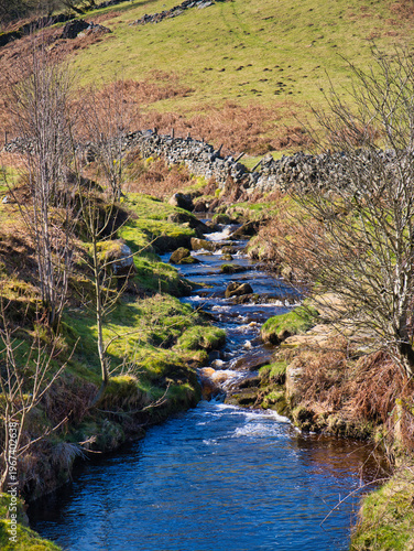 A clear winter stream winds through grassy fields and dry stone walls in rural Yorkshire UK, with bright light and crisp countryside detail