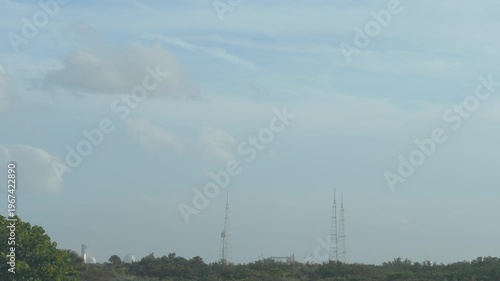 A rocket launches from a coastal space center with communication towers and infrastructure visible under a cloudy sky. Bright engine flames rise above trees and buildings as the spacecraft lifts off