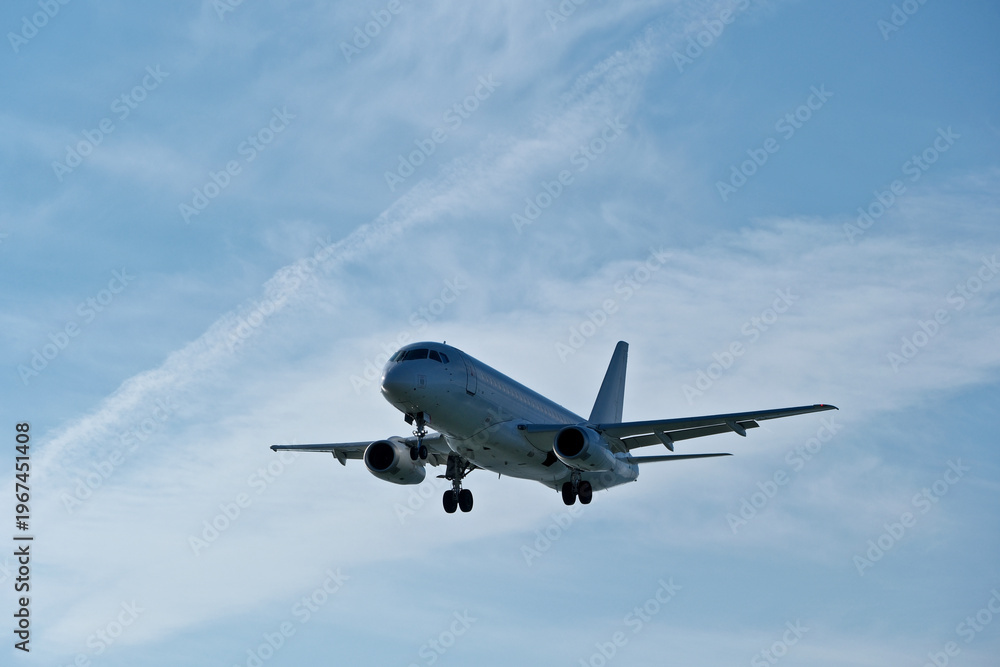 Obraz premium White passenger plane on final approach on a sunny day with blue sky, light clouds and beautiful backlighting. Sunlit white airplane landing against a blue sky with wispy clouds, backlit by bright sun
