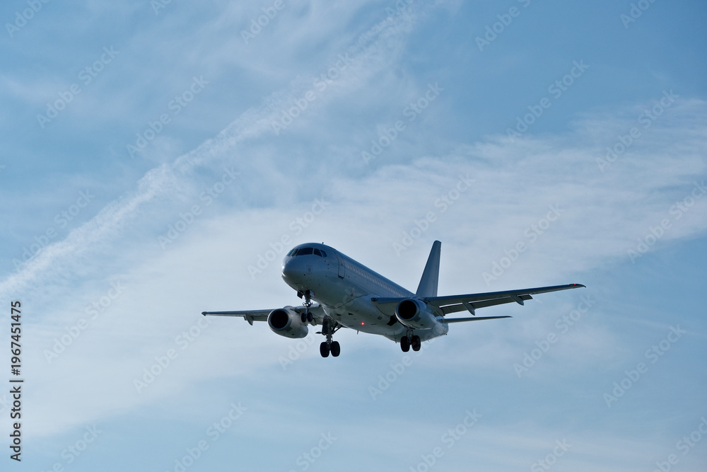 Fototapeta premium White passenger plane on final approach on a sunny day with blue sky, light clouds and beautiful backlighting. Sunlit white airplane landing against a blue sky with wispy clouds, backlit by bright sun