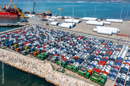 An aerial view of a large port where hundreds of new cars are lined up in orderly rows for export. Cargo ships, cranes, trucks and industrial equipment, global logistics, automotive trade.