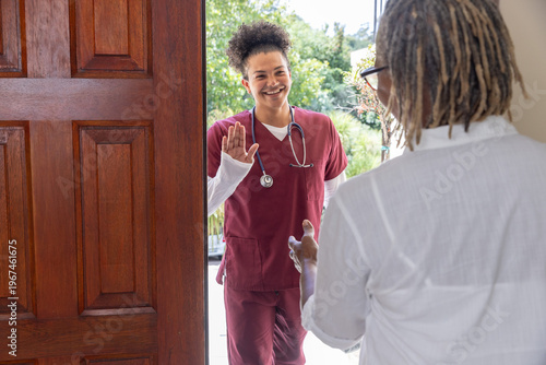 Mid adult nurse in maroon scrubs with stethoscope greeting senior African American woman at doorway