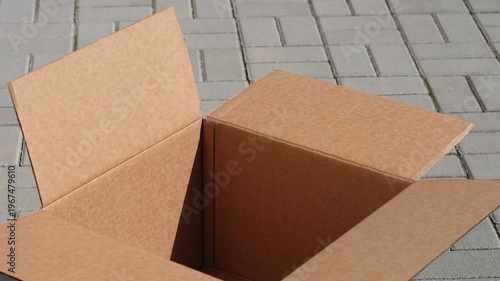 An empty open cardboard box sitting on a grey paved outdoor surface with its flaps fluttering in the wind. Close up.