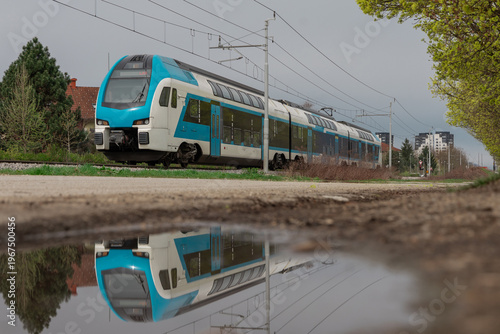 Modern passenger train traveling on railway tracks with overhead electric lines, reflected in a puddle in the foreground, capturing urban transport, motion, and symmetry in a calm suburban setting.