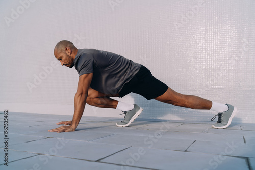 Side view of athlete doing deep lunge stretch on outdoor floor, morning fitness and mobility workout, urban training environment, healthy man exercising in sunlight, strength and balance concept.