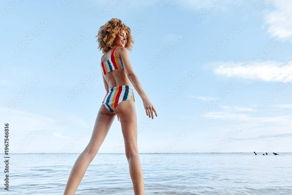 Naklejka premium Woman with curly hair wearing colorful striped swimsuit standing on the shore with water and clear sky. Concept of summer lifestyle, beach, and outdoor relaxation.