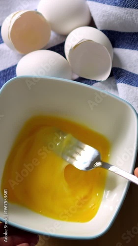 Toronto, Canada, January 12, 2026: Fresh eggs cracked whisked in bowl yellow yolk stirred with fork in white ceramic bowl beside cracked shells on striped towel home cook prepping omelette gentle