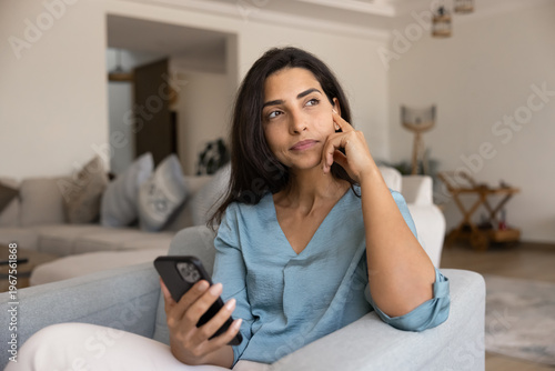 Принти на полотні Brazilian woman waits for call seated on armchair with smartphone at home, looks