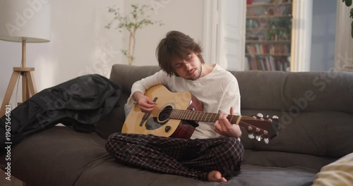 A young musician sits cross-legged on his sofa, lost in music as he strums his guitar softly in a warm, inviting space