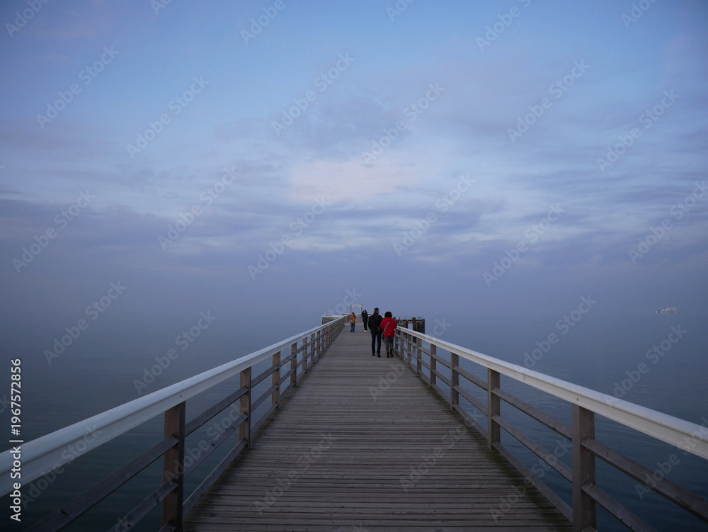 Fototapeta premium A wooden pier extends out over calm water towards a hazy horizon. Silhouetted figures walk along the pier under a pale, cloudy sky.