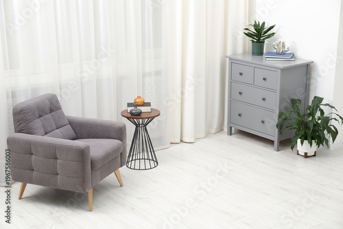 Stylish armchair, side table, chest of drawers, decor and houseplants near window indoors. Interior design