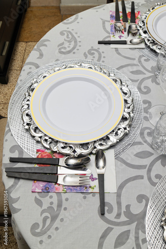 Dining table set for eight with elegant white tablecloth, silver charger plates, glassware and organized cutlery for formal home dinner