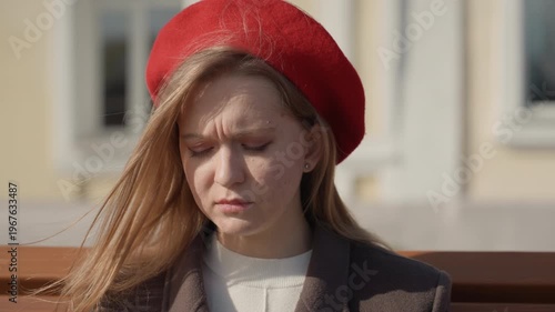 White woman on bench receiving call, red beret fluttering, furrowed brow and worried expression, sunlight highlights hair, urban facade background, moment of decision and resolve, closeup portrait