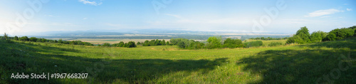 A beautiful summer panoramic landscape with a meadow on a mountain slope. A vast mountain valley is visible in the distance. Kuban nature. Caucasus foothills. Krasnodar Krai, Russia. Wide panorama.