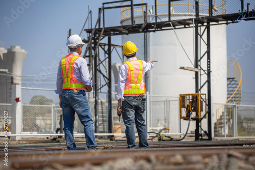 Engineers in safety vests and hardhats discussing project plan at industrial factory site, Back view of industrial inspectors standing on railway track pointing at storage tank area