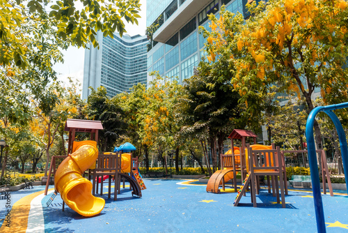 Colorful modern children's playground in a public park surrounded by city skyscrapers and yellow flowering trees.