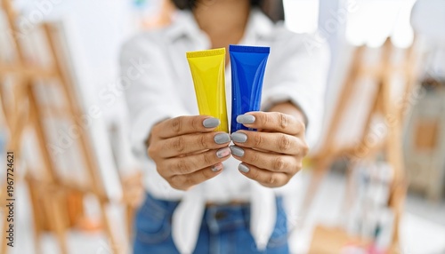 Artist holding yellow and blue paint tubes in a studio.