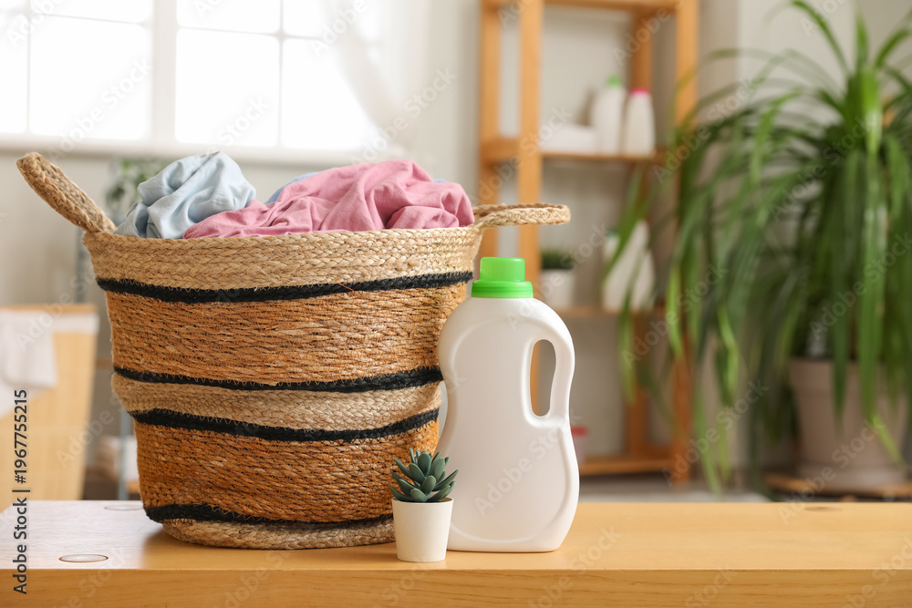 © Pixel-Shot - Wicker basket with laundry, houseplant and bottle of detergent on wooden table in room