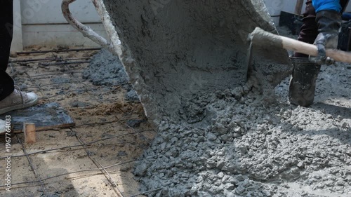 An industrial construction worker with a shovel handles heavy sand and cement at an outdoor building site filled with steel and stone equipment