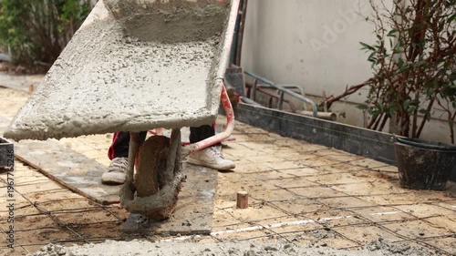 An old metal wheelbarrow stands near a weathered brick wall and wooden building at a garden construction site