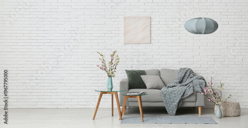 Sofa with coffee tables and vases of blooming branches near white brick wall in living room