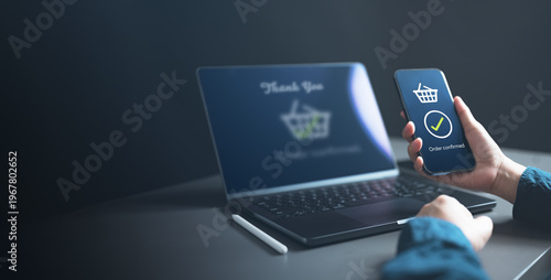 Fotografie A person holding a smartphone showing an order confirmed screen beside a laptop, symbolizing successful online shopping, digital transaction completion, and e-commerce payment confirmation