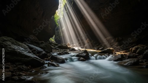 Dramatic sunbeams pierce through a dark cave opening, illuminating a flowing river with smooth, long-exposure water and rugged rock formations.