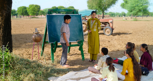 Indian happy young adult gen z female do work talk to villager girl boy sitting on mat rural area. teen male child writing study look board enjoy desi life open day class field ground place