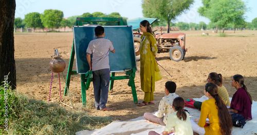Indian happy young adult gen z female do work talk to villager girl boy sitting on mat rural area. teen male child writing study look board enjoy desi life open day class field ground place