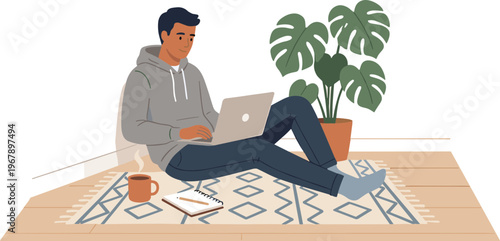 A man sits on a rug working on laptop with plant nearby