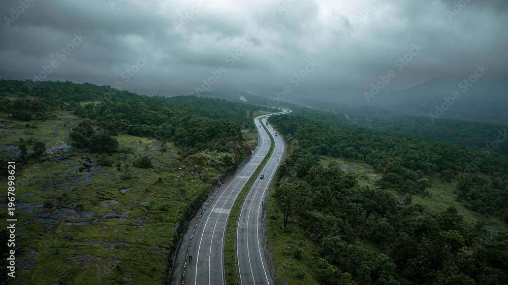 Fototapeta premium A dramatic aerial view of a highway in India winding through lush green hills and dense forest under a cloudy sky. Vehicles travel along the smooth, curving road, highlighting modern infrastructure.