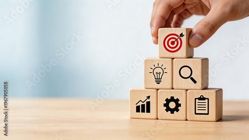 Hand placing wooden block with target icon on pyramid of blocks featuring business symbols for strategy planning growth innovation analysis and management concepts on desk
