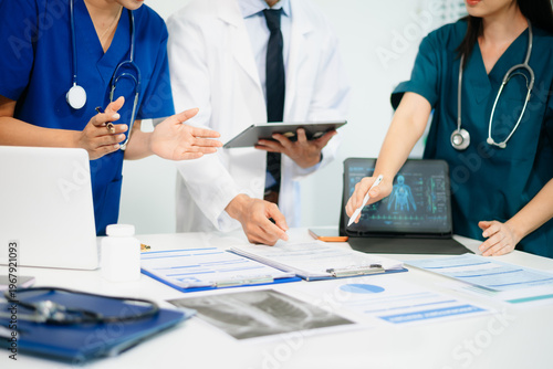 Medical team having a meeting with doctors in white lab coats and surgical scrubs seated at a table discussing a patients working online using computers