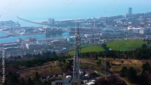 Sideways Aerial View of TV Radio Transmitter Tower High Above Swansea City with Marina in Distance. 5G Infrastructure for Smart Phones and Connectivity.