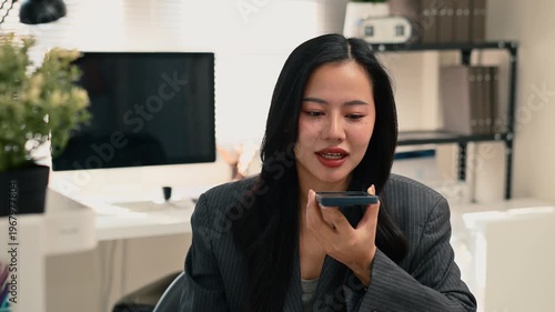 Professional woman using voice assistant chatbot voice on smartphone while working on laptop in modern office.